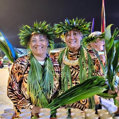 Deux femmes en tenue traditionnelle polynésienne portant couronnes de feuilles tressées et colliers de fleurs lors d'une soirée culturelle — gardiennes de la transmission orale māʻohi